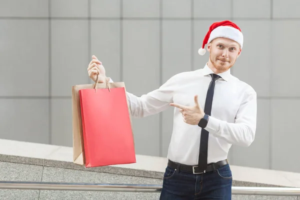 Svart fredag på vintern. Lycka röda huvud affärsman i santa hatt, håller kassar på händer, peka finger och titta på kameran. — Stockfoto
