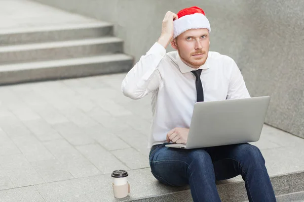 Práce v přírodě, sedí na zemi. Mladých dospělých zázvor vousatých podnikatel v santa hat, práce na pc a snění. — Stock fotografie