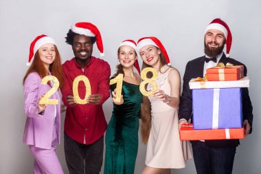 Sayı 2018 tutarak ve dişlek gülümseyerek kameraya bakarak genç yetişkin arkadaşlarım. Bir yakın duran ve birçok hediye kutusu tutan adam sakallı. Kapalı atış, gri arka plan