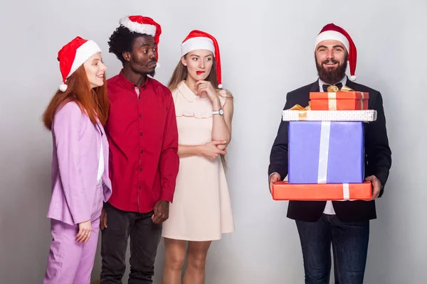Barbu homme bien habillé dans la casquette de Noël, tenant de nombreux coffret cadeau. Ses amis se tiennent près, regardent la boîte et devinent ce qu'il y a à l'intérieur. Plan studio, fond gris — Photo