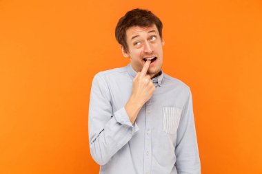 Something is stuck between teeth. Funny young adult business man touching finger teeth and looking up. Studio shot, orange background clipart