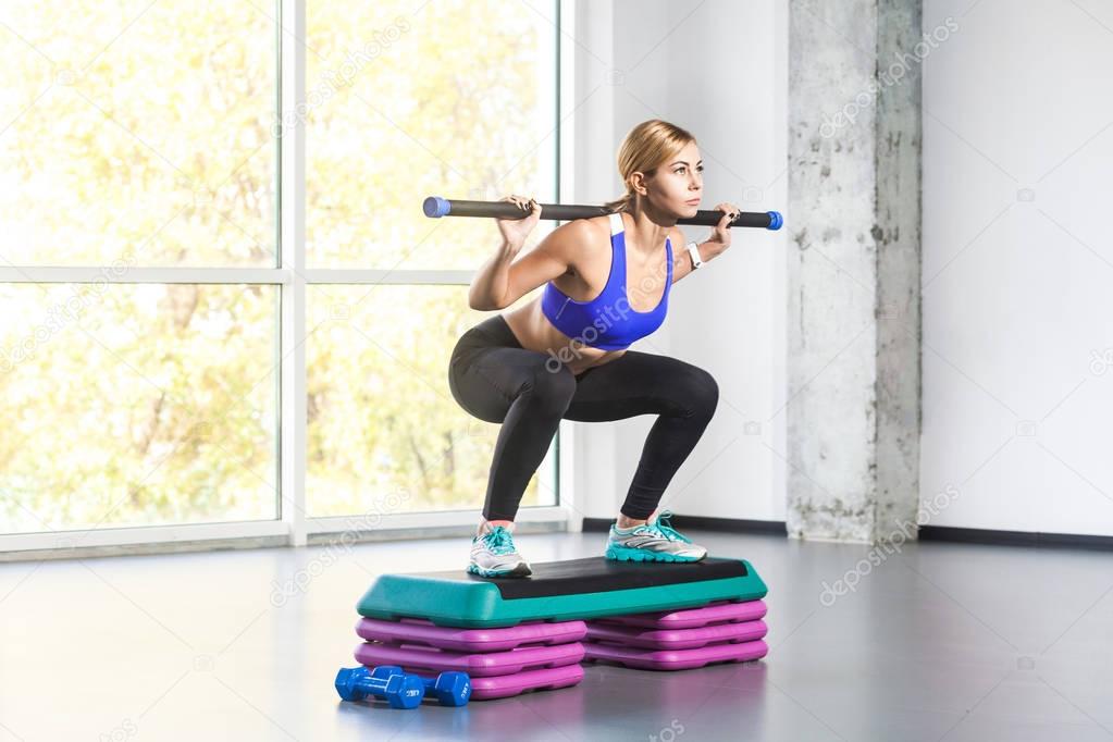 Blonde woman doing squats on step platform with barbell bar 