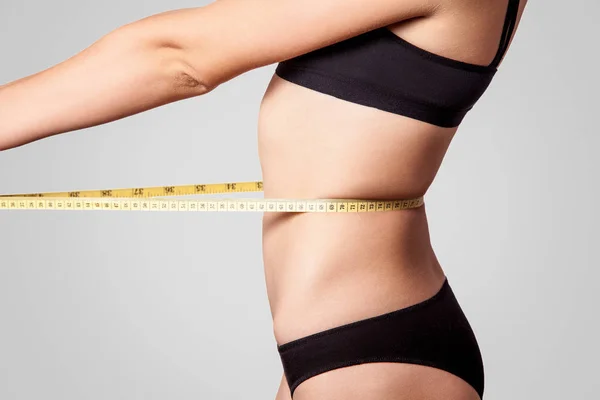 明るい灰色の背景 ダイエットと痩身のコンセプトにメジャー テープでウエストを測定黒下着でスリムな若い女性の側面図 — ストック写真