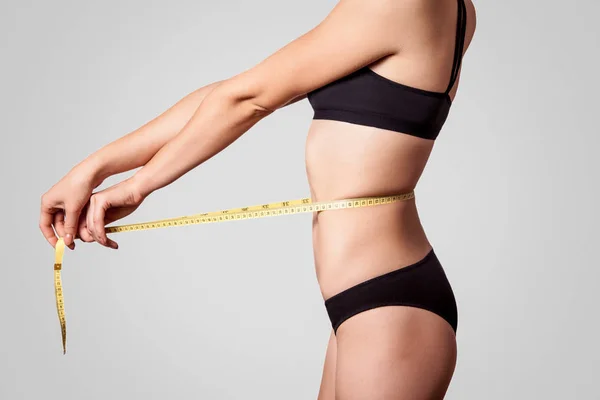 明るい灰色の背景 ダイエットと痩身のコンセプトにメジャー テープでウエストを測定黒下着でスリムな若い女性の側面図 — ストック写真
