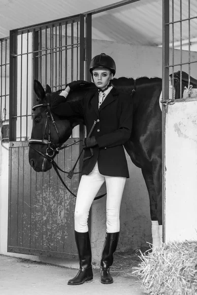 attractive woman wearing jockey equipment posing near big black horse in stable, sport and fashion concept