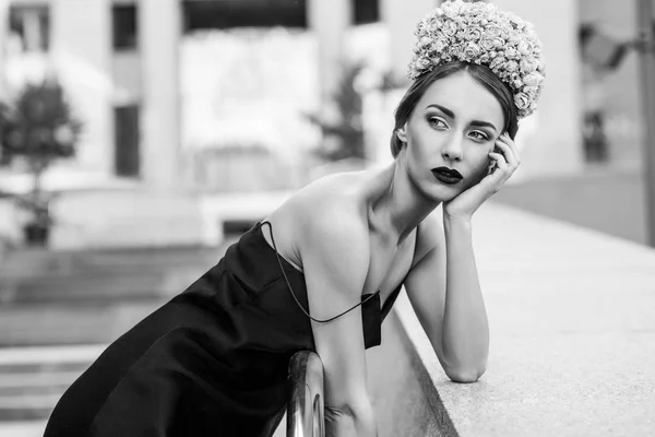 花の冠と階段の上の金属の手すりにもたれて黒のドレスを着て豪華な女性 — ストック写真