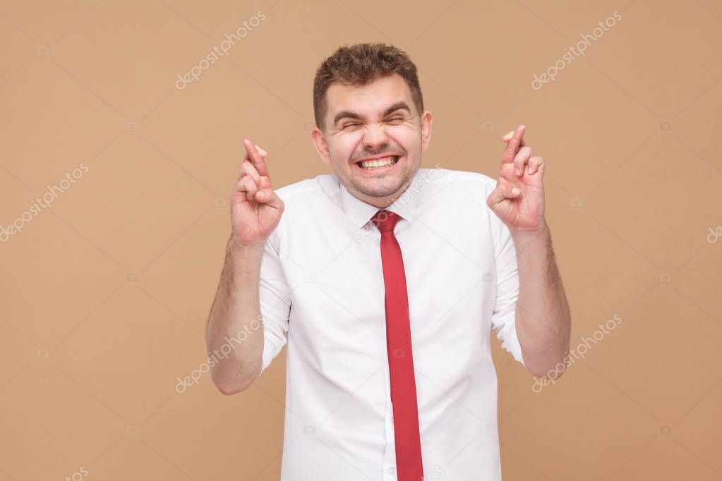 smiling businessman with closed eyes crossed fingers on light brown background , hope and wish concept 