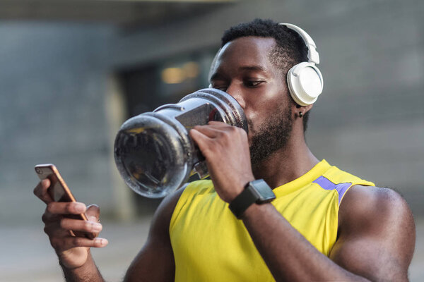 strong man drinking water from bottle after hard workout and looking at screen of smartphone in hand 