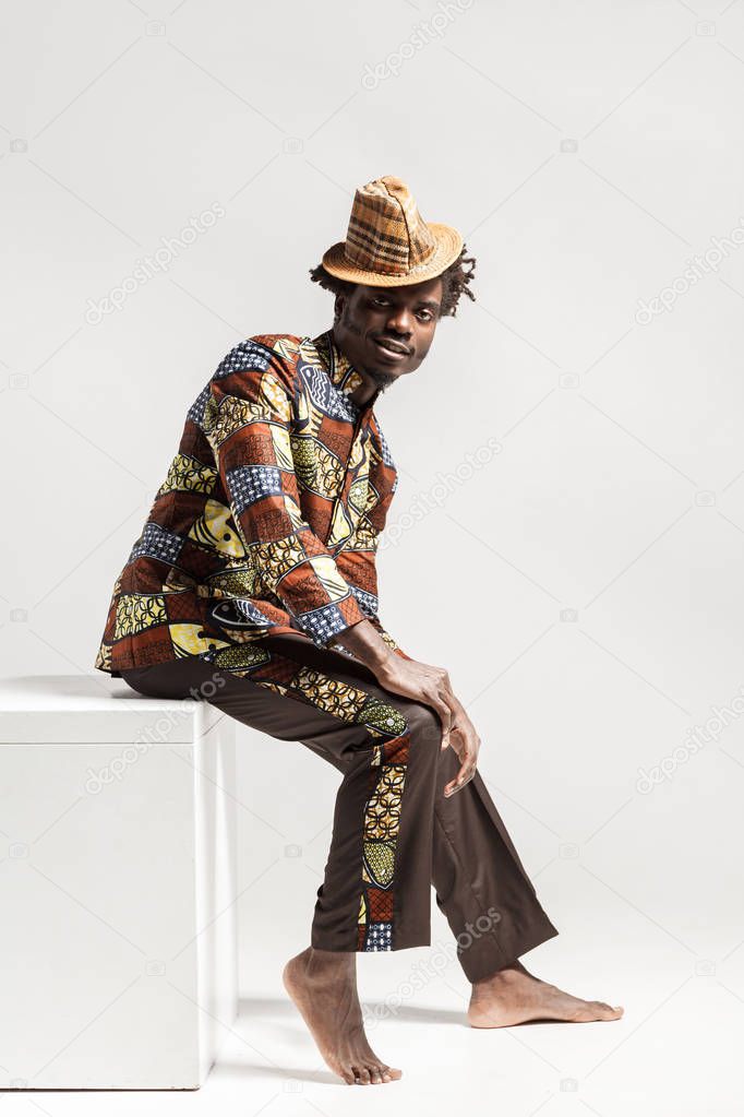 handsome african american man wearing costume in national Kongo style sitting on cube isolated on white background