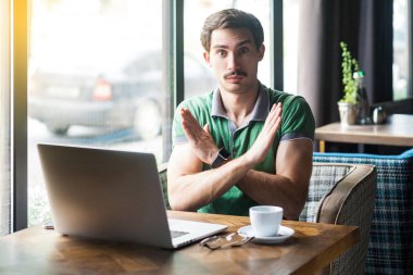 Young serious businessman in green t-shirt looking at camera and showing closed sign with hands while sitting at table with laptop in cafe, business and freelancing concept