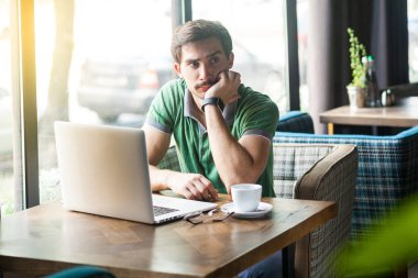 Young thoughtful quizzical businessman in green t-shirt holding hand on chin and looking away while sitting at table with laptop in cafe, business and freelancing concept 
