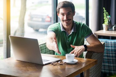 Young happy businessman in green t-shirt looking at camera and giving hand to greeting while sitting at table with laptop in cafe, business and freelancing concept