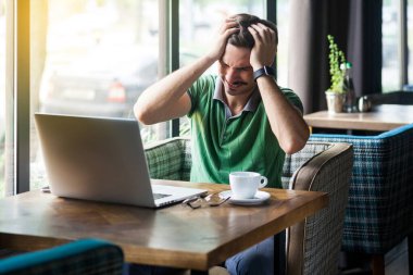 Young businessman in green t-shirt holding his painful head while sitting at table with laptop in cafe, business and freelancing concept