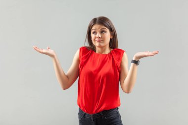 confused beautiful brunette young woman in red t-shirt standing with raised arms and looking away on gray background