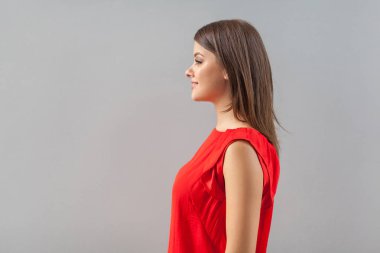 side view of happy beautiful brunette young woman in red t-shirt standing and looking forward with toothy smile and happiness on gray background