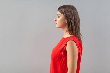 side view of calm beautiful brunette young woman in red t-shirt standing and looking forward with serious face on gray background