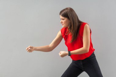 side view of serious beautiful brunette young woman in red t-shirt standing with fists and try to pulling on gray background