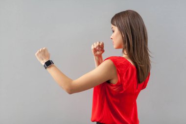 side view of serious beautiful brunette young woman in red t-shirt standing with boxing fists and ready to attack on gray background