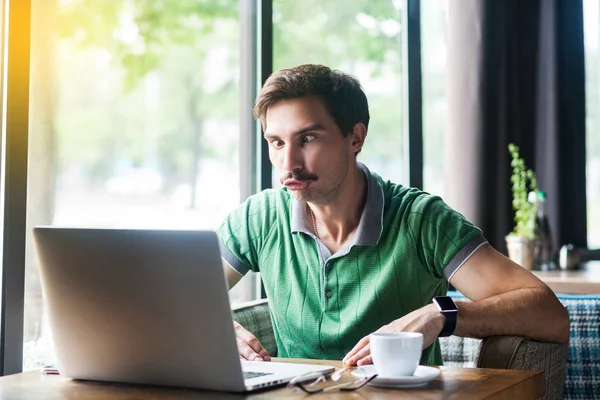 Young funny businessman in green t-shirt looking at laptop display with stupid crazy face and crossed eyes while sitting at table in cafe, business and freelancing concept