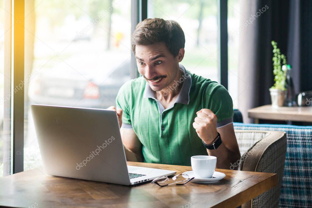 Young happy surprised businessman in green t-shirt looking at laptop screen and rejoicing success while sitting at table in cafe, business and freelancing concept 