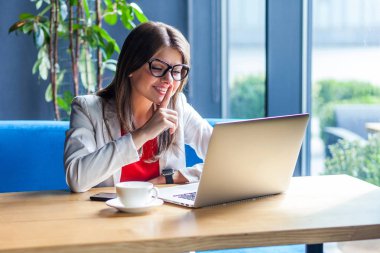 toothy smiley beautiful stylish young woman in glasses looking at laptop screen and showing secret sign during video call while sitting at table in cafe