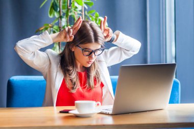 angry beautiful stylish brunette young woman in glasses looking at laptop screen and showing cow horns gesture with anger face while sitting at table in cafe