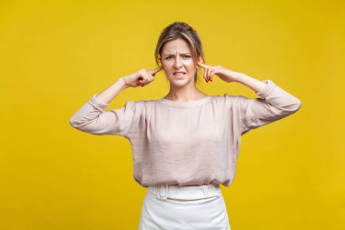 I don't want to hear you. Portrait of annoyed young woman with fair hair in casual blouse standing, closing ears with fingers and looking at camera. indoor studio shot isolated on yellow background clipart