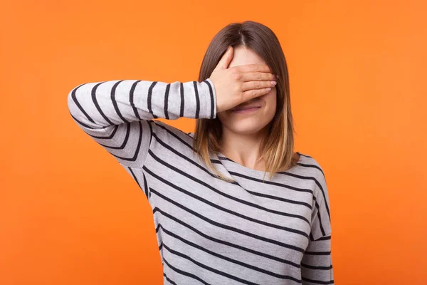 Don't want to look at this. Portrait of young scared woman with brown hair in long sleeve shirt standing, covering eyes with hand, refusing to watch. indoor studio shot isolated on orange background