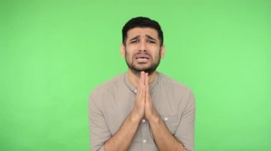 Tişörtünde kılları olan, dua ederken ellerini tokuşturan, neredeyse ağlayan, lütfen dileyen, merhamet dileyen zavallı esmer adam. Stüdyo çekimi, yeşil arkaplan, krom anahtar