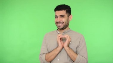 Nazik yürekli, tişörtlü esmer adam kalp hareketi yapıyor ve sevgililer gününde romantik duygular ifade ederek seni seviyorum diyor. Stüdyo içi çekim, yeşil arka plan, krom anahtar.