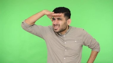 Tişörtünde kıl olan kararlı esmer adam avucunu gözlerine bastırıyor ve kameraya bakarken dikkatlice uzaklığa bakıyor gülümsüyor. Stüdyo içi çekim, yeşil arka plan, krom anahtar.