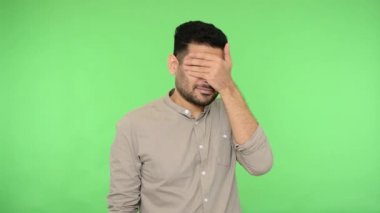 Meraklı esmer adam, tişörtünde tüy yumağı, yüzünü elleriyle kapatıyor ve parmaklarının arasından garip, şaşırmış bir ifadeyle, utanç verici bir sırrı dikizliyor. Stüdyo içi çekim, yeşil arka plan, krom anahtar.