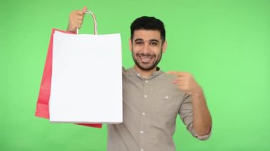 Tişörtlü, mutlu bir erkek alıcı elindeki alışveriş torbalarını işaret ediyor ve iyi niyet göstergesi olarak paketler halinde iyi şeyler öneriyor. Stüdyo içi çekim, yeşil arka plan, krom anahtar.