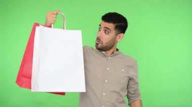Tişörtlü mutlu erkek alıcı, alışveriş çantalarını işaret ediyor ve şaşırmış görünüyor, satın almaktan memnun görünüyor, başparmaklarını kaldırıyor, jest gibi. İç mekan stüdyo çekimi, kroma anahtarı.