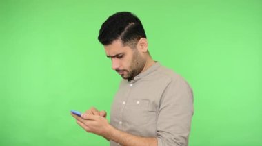 Tişörtünde kıvrımları olan esmer, ciddi bir adam sosyal medyada geziniyor ya da cep telefonundan mesaj okuyor, mobil servisleri kullanarak internetten alışveriş yapıyor. Stüdyo içi çekim, yeşil arka plan, krom anahtar.