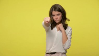 Saldırgan esmer kadın, bluz giyen esmer kadın boks yapıyor, jest yapıyor, kameranın önünde kararlı bir ifadeyle yumruk atıyor, nefsi müdafaa yapıyor. Stüdyo çekimi, sarı arkaplan