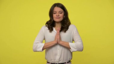 Sarışın, bluzlu barışçıl bir kadın elleriyle dua ediyor, düşünceli yoga yapıyor, sakin bir ifade takınıyor. Sarı arka planda izole edilmiş kapalı stüdyo çekimi