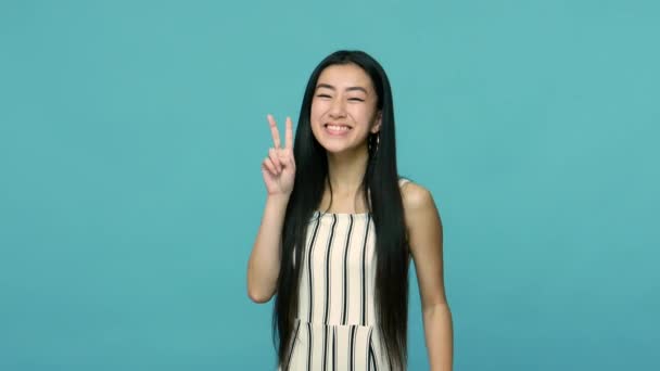 乐观的亚洲女人 有着长长的笔直的黑发 有双V字 有胜利或和平的手势 大大的微笑 庆祝成功 室内拍摄 蓝色背景隔离 — 图库视频影像