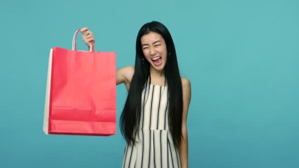 パッケージを指して はい勝利ジェスチャーを示す長い黒い髪を持つ非常に幸せなアジアの女性は 購入に満足し ショッピングを楽しんで 青い背景に隔離された屋内スタジオで — ストック動画