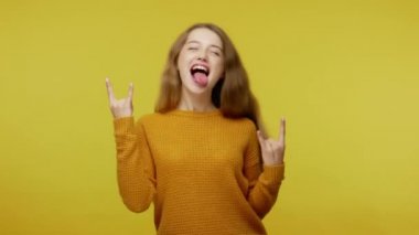 İtaatsiz, kahverengi saçlı, kazaklı hoş bir kız. Rock and roll el hareketi yapıyor. Havalı bir işaret veriyor ve dilini gösteriyor. Stüdyo çekimi sarı arkaplanda izole edildi