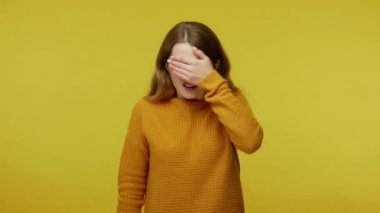 No, I don't want to look! Timid frightened young woman in pullover covering eyes with hand and showing stop gesture, confused shy scared to watch. indoor studio shot isolated on yellow background