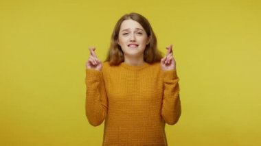 Cute girl with brown hair in pullover hoping for fortune success, making wish and holding fingers crossed for good luck, waiting her dreams come true. indoor studio shot isolated on yellow background