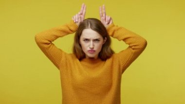Irritated girl in pullover showing bull horns with fingers on head and making threatening grimace to camera, her look expressing anger and danger. indoor studio shot isolated on yellow background