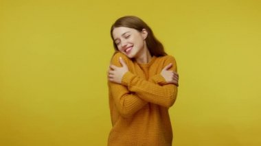 Beautiful lovely happy girl with brown hair in pullover embracing herself and smiling with dreamy satisfied expression, love yourself concept. indoor studio shot isolated on yellow background
