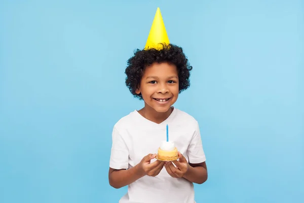 En liten søt, lykkelig gutt med en festlig kjegle på hodet som holder kake – stockfoto