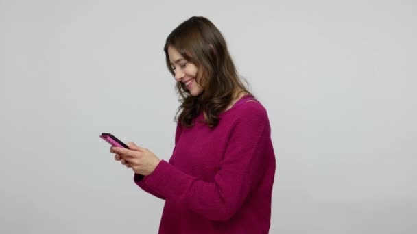 Happy charming brunette woman in pullover using mobile phone, typing message on social network and laughing — Stockvideo