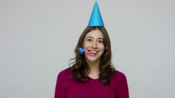 Humorística mujer morena feliz con cono divertido en la cabeza soplando cuerno de fiesta, felicitando en aniversario de cumpleaños — Vídeos de Stock