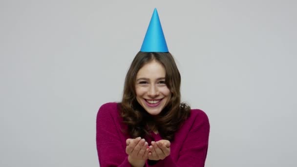Happy birthday! Fascinating brunette woman with funny party cone on head blowing glitters, scattering confetti — 图库视频影像