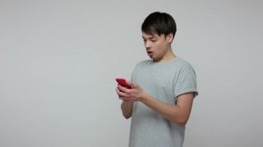 Mobile internet usage. Surprised guy in t-shirt scrolling pictures on social networks, reading shocking news, watching blog with interest and amazement. indoor studio shot isolated on gray background