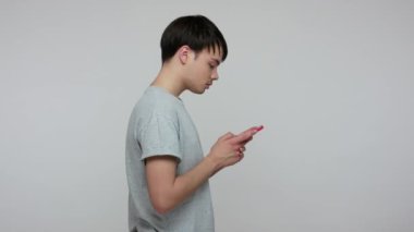 Mobile communication. Side view of young brunette guy in T-shirt typing message on cell phone, chatting in social network using online application. indoor studio shot isolated on gray background